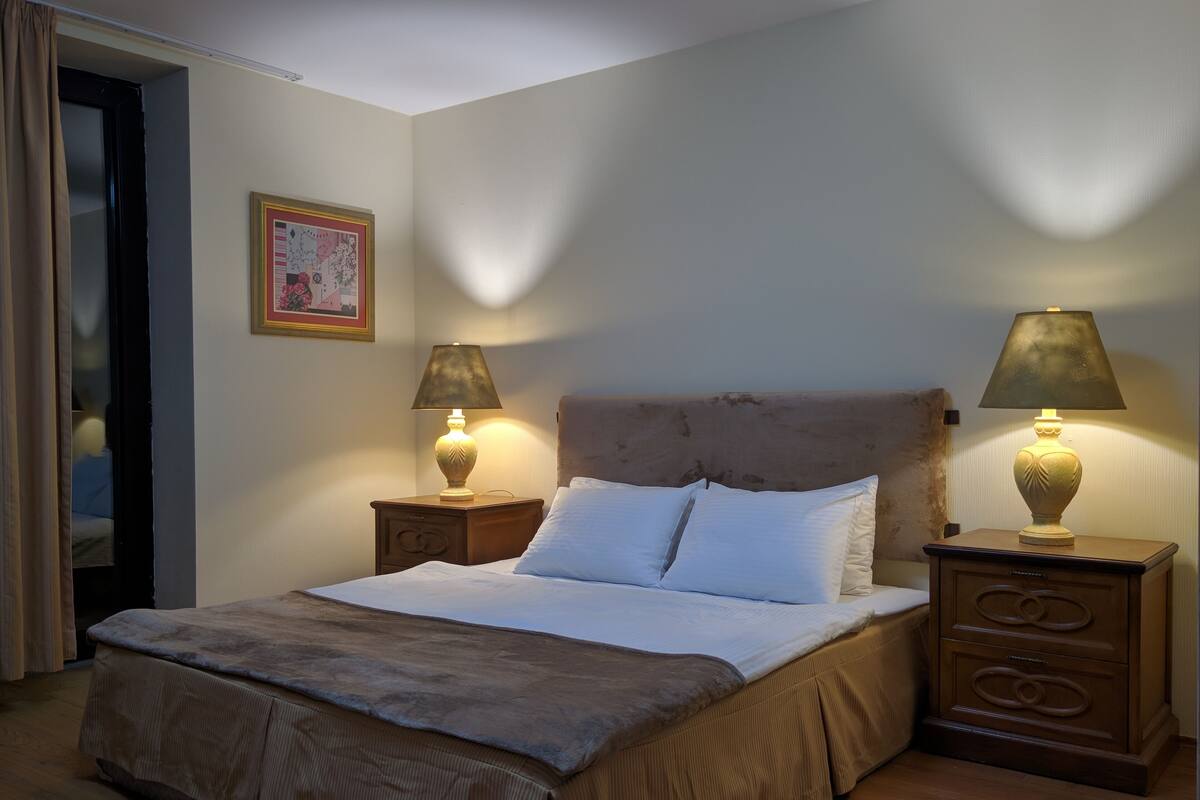 Ein ordentlich bezogenes Bett mit wei ßen Leinen, zwei Nachttischlampen, ein gerahmtes Bild an der Wand und ein Nachttisch mit Schubladen.