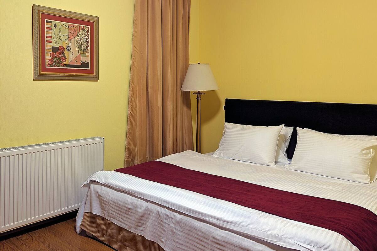 Ein Bett mit weißer und purpurroter Bettwäsche, ein schwarzer Kopfteil, ein Heizkörper, eine Lampe und ein gerahmtes Bild an der Wand.