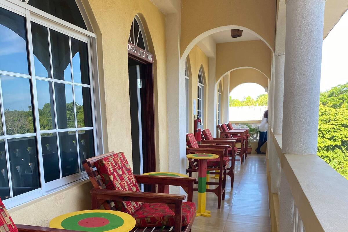 A covered outdoor seating area with wooden chairs and tables, a tiled floor, and a view of greenery.