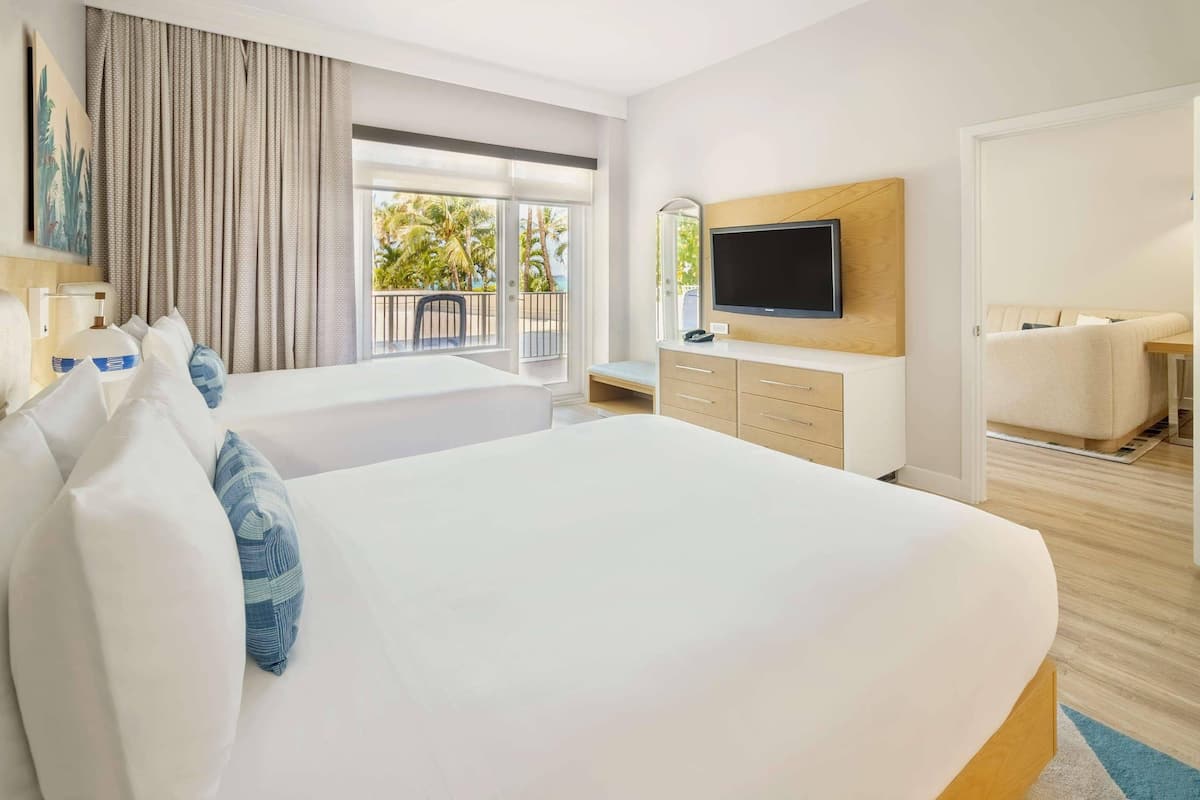 A hotel room with a large bed, a TV, a desk, and a balcony with a view of palm trees.