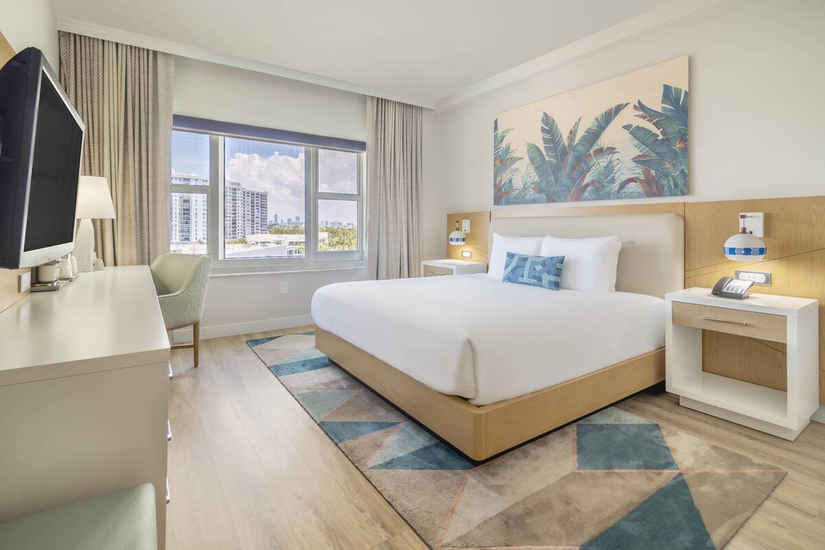A hotel room with a large bed, a desk with a TV, a chair, a nightstand with a phone, and a window with a city view.