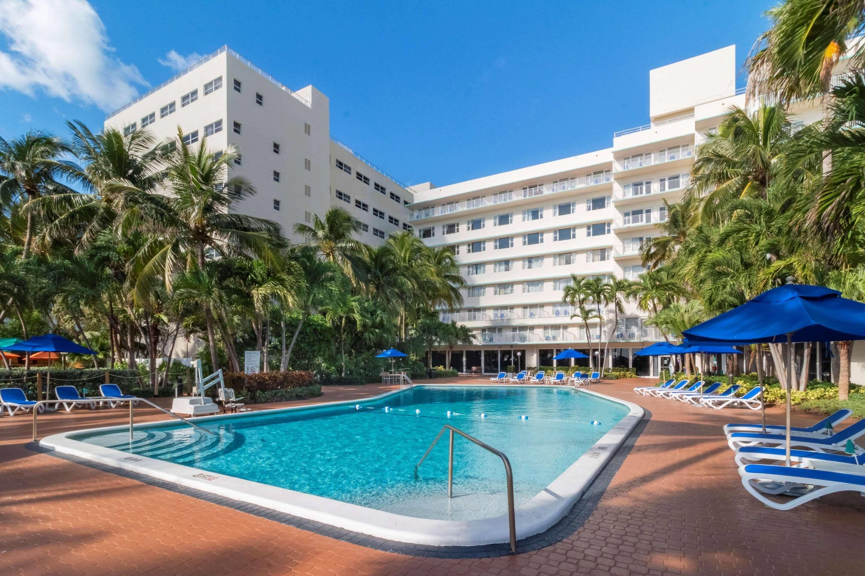 A swimming pool with blue umbrellas and lounge chairs, surrounded by palm trees and a multi-story hotel building.