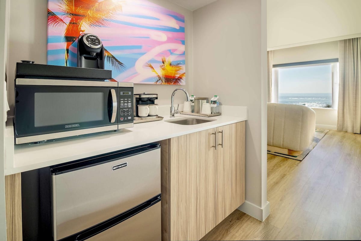 A compact kitchen with a microwave, coffee maker, and sink. A painting of a tropical scene adorns the wall.