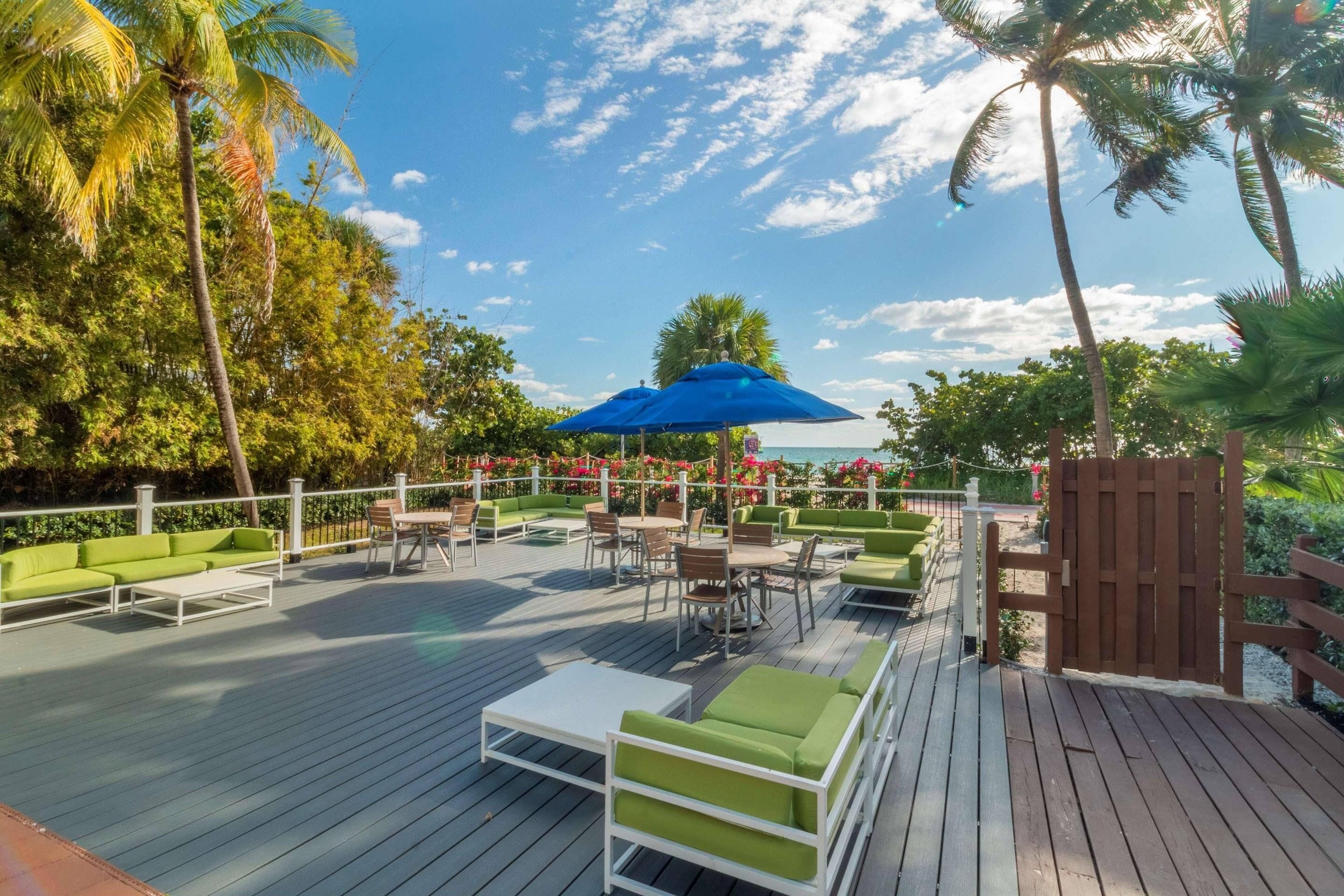 Een houten terras met buitenmeubels, waaronder groene banken en tafels, onder een blauwe parasol.