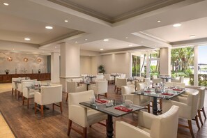 A spacious dining area with multiple tables and chairs, large windows with a view of palm trees, and a decorative wall with a fountain. - Radisson Resort Miami Beach  (Miami Beach)