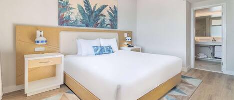 Una habitación de hotel moderna con una cama grande, un cabecero de madera, una mesita de noche y una lámpara de pared, junto con un cuadro de hojas tropicales.