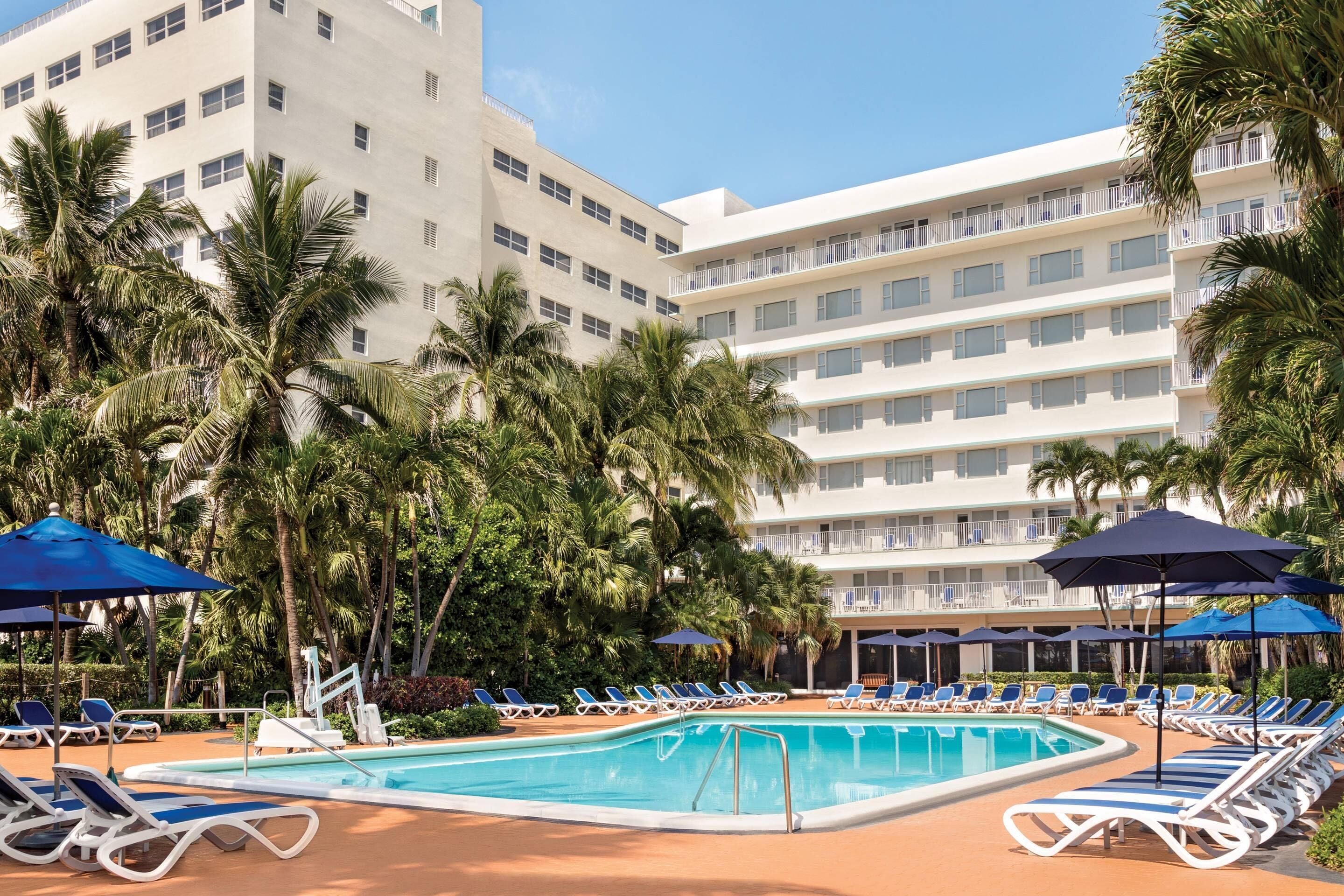 A pool area with lounge chairs and umbrellas, surrounded by palm trees and a multi-story hotel building.