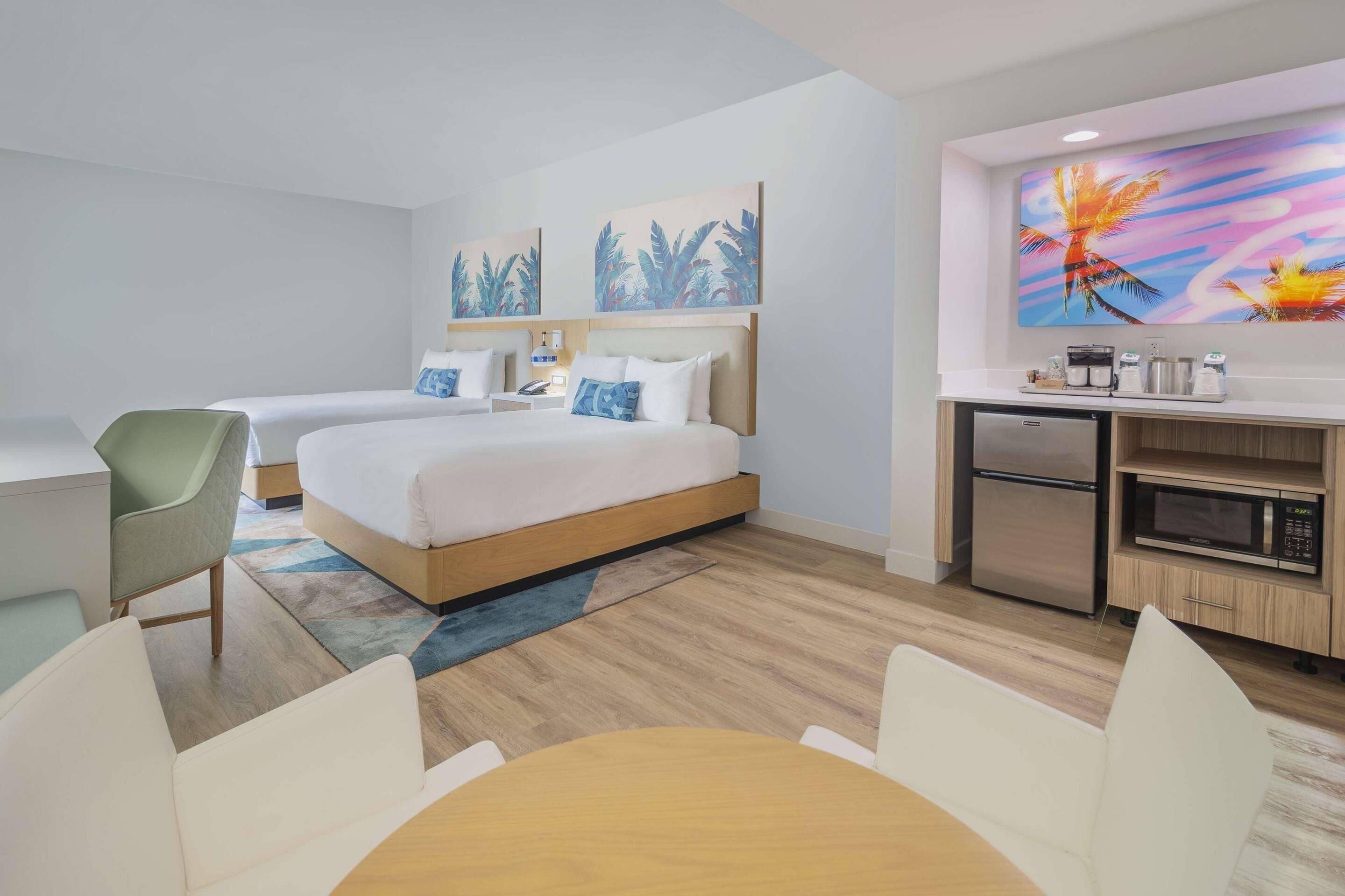 A hotel room with a bed, a desk, a chair, a dining table, a kitchenette, and artwork on the walls.