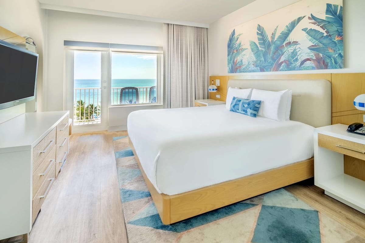 A hotel room with a large bed, a TV, a desk, and a view of the ocean.