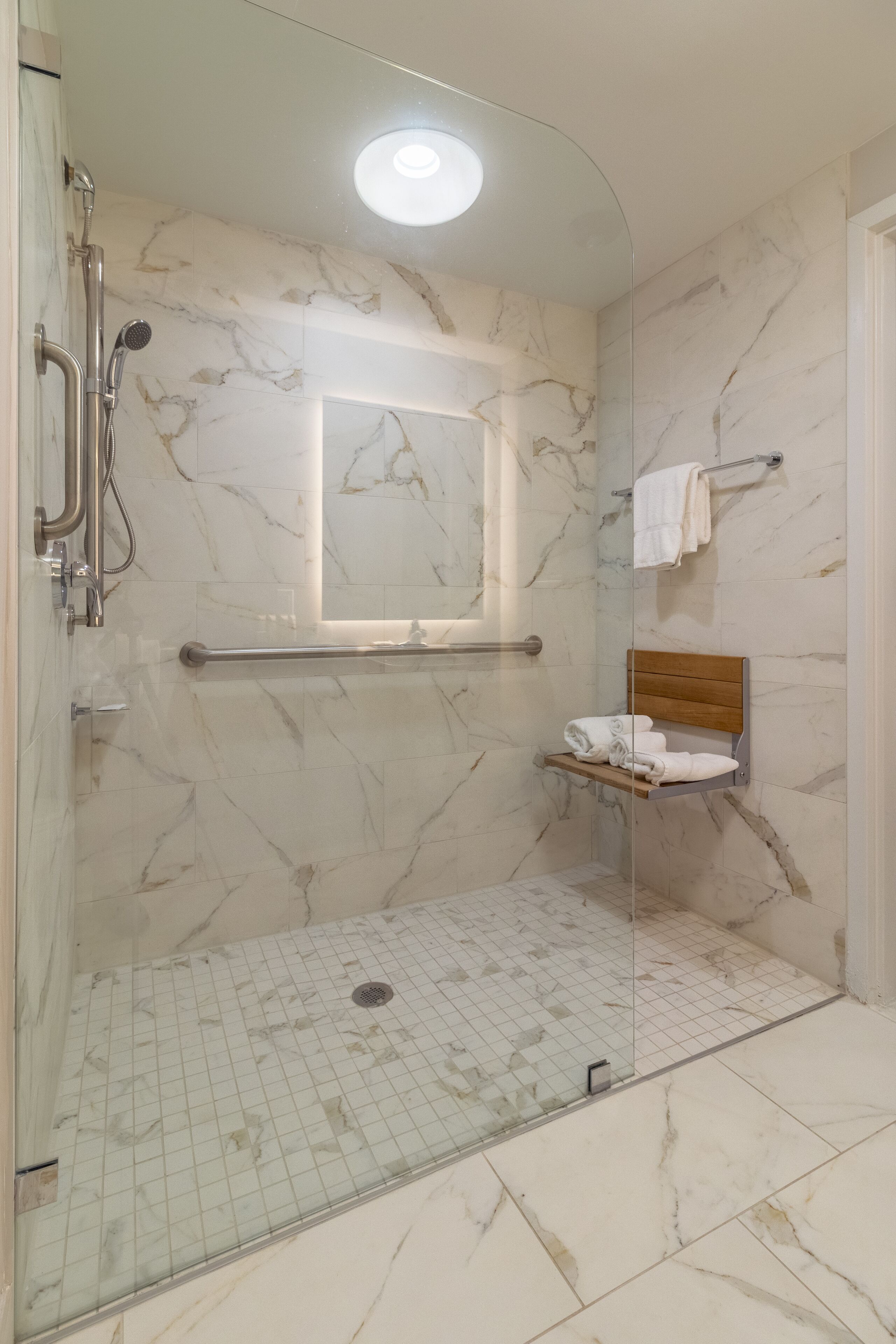 Un baño moderno con ducha de vidrio, paredes de mármol, repisa de madera con toallas y pasamanos.