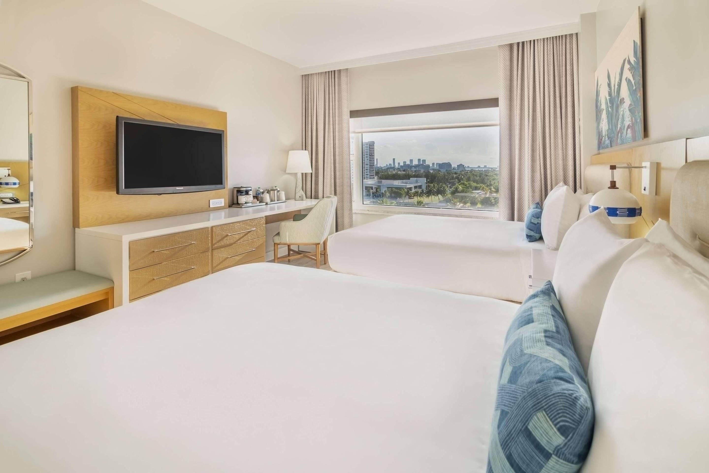 A hotel room with a flat-screen TV, a desk with a lamp, two beds, a window with a city view, and a large painting on the wall.