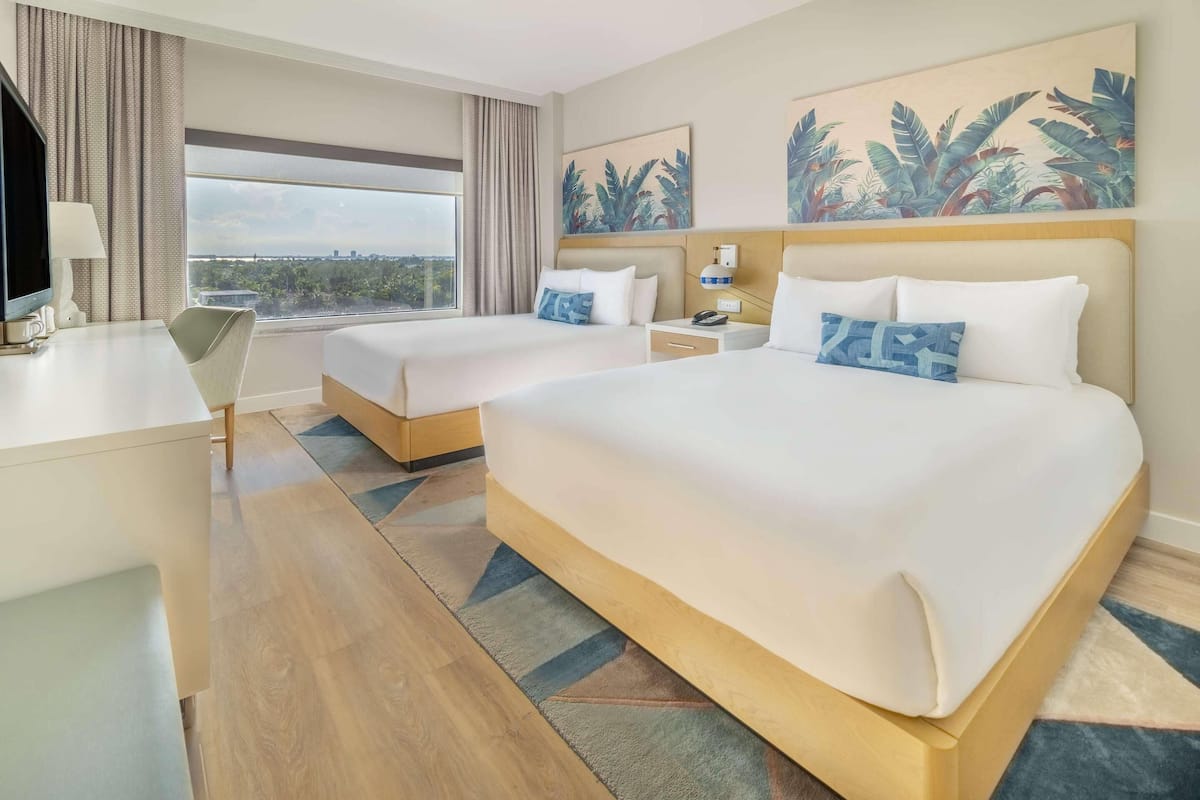 A hotel room with two beds, a desk, a chair, and a large window offering a city view.