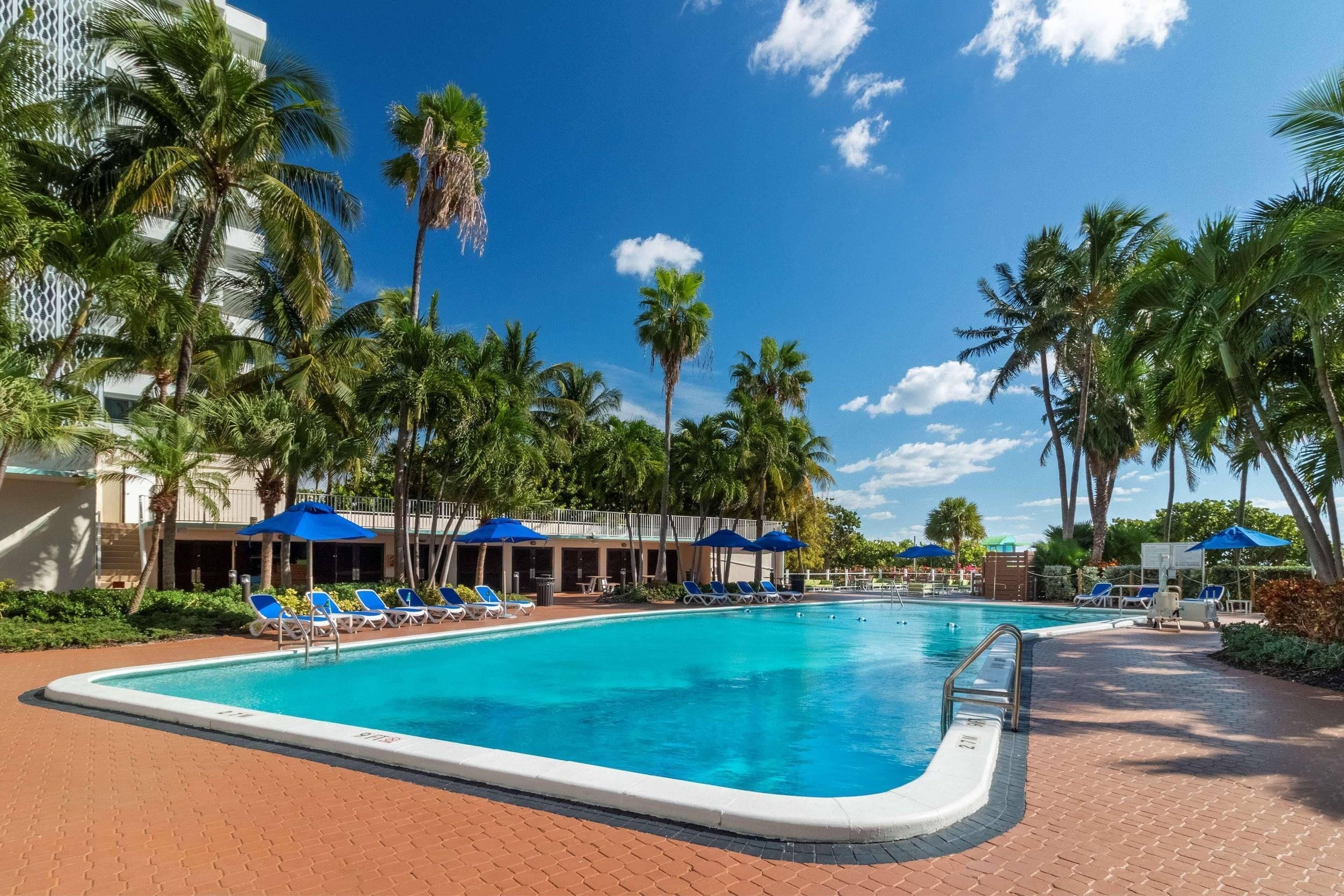 A swimming pool with lounge chairs and umbrellas, surrounded by palm trees and a building with a white facade.