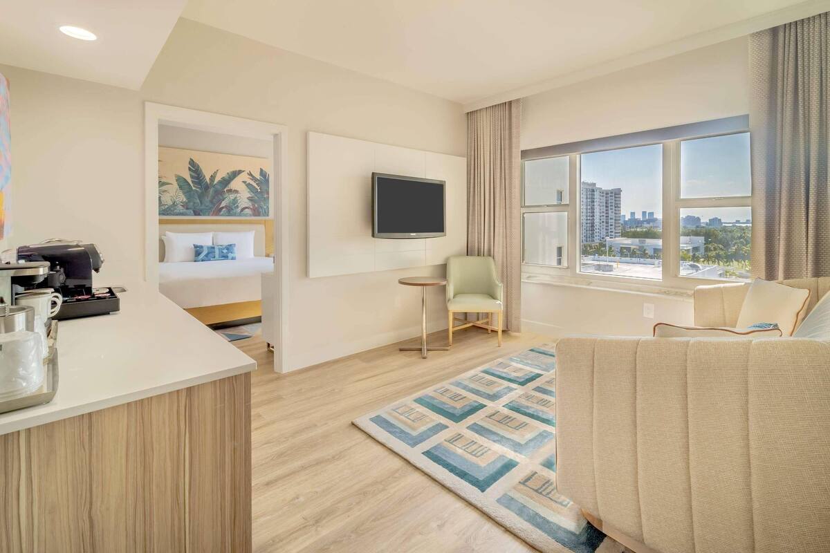 A hotel room with a bed, a TV mounted on the wall, a small table with a chair, and a view of the city through large windows.