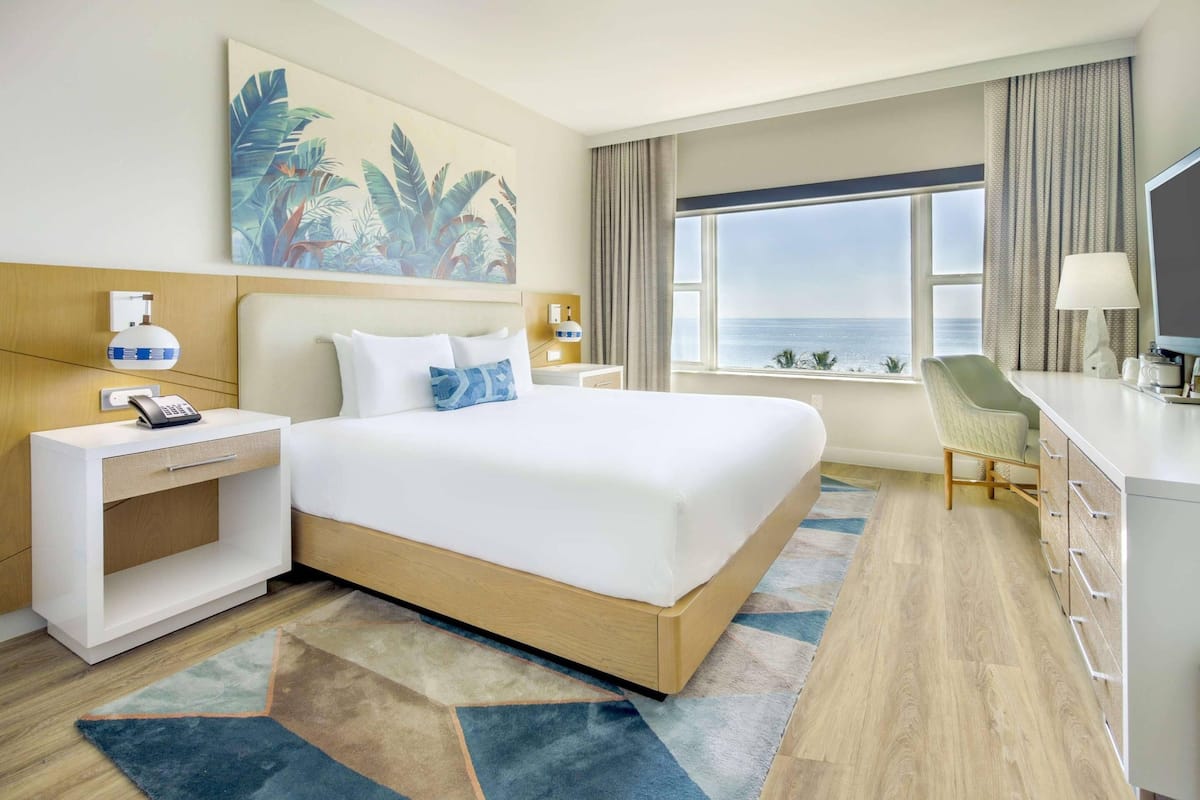 A hotel room with a large bed, a desk, a chair, a nightstand, a phone, a lamp, a painting, a window with a view, and a rug.