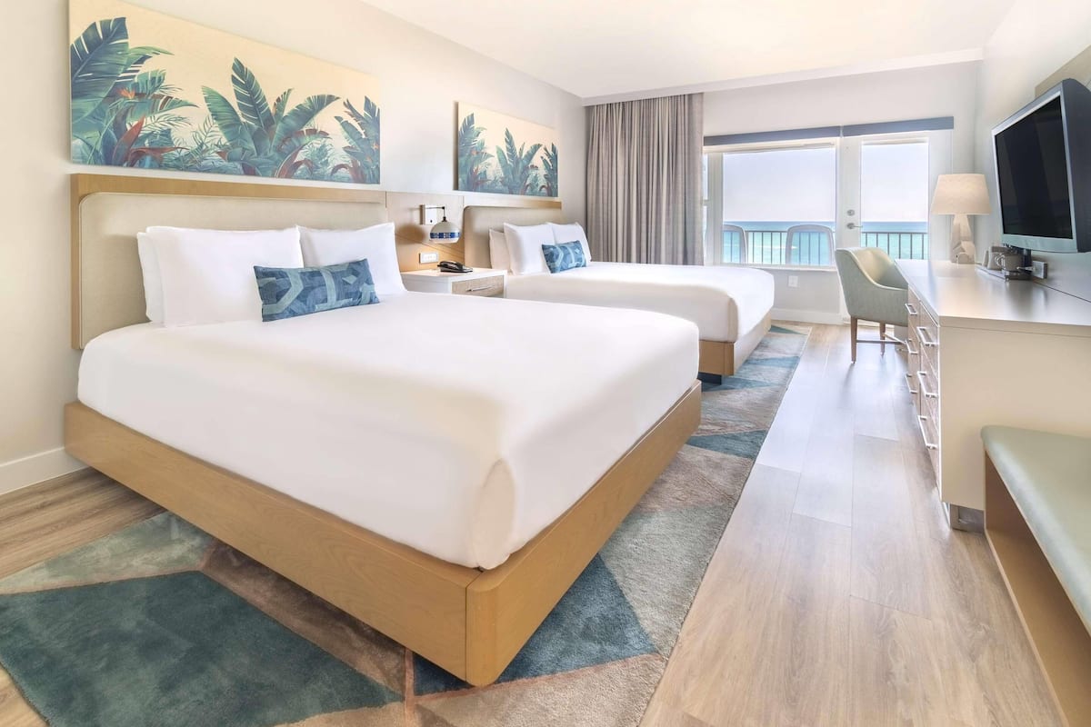 A hotel room with two beds, a desk, a chair, a TV, and a balcony with a view.