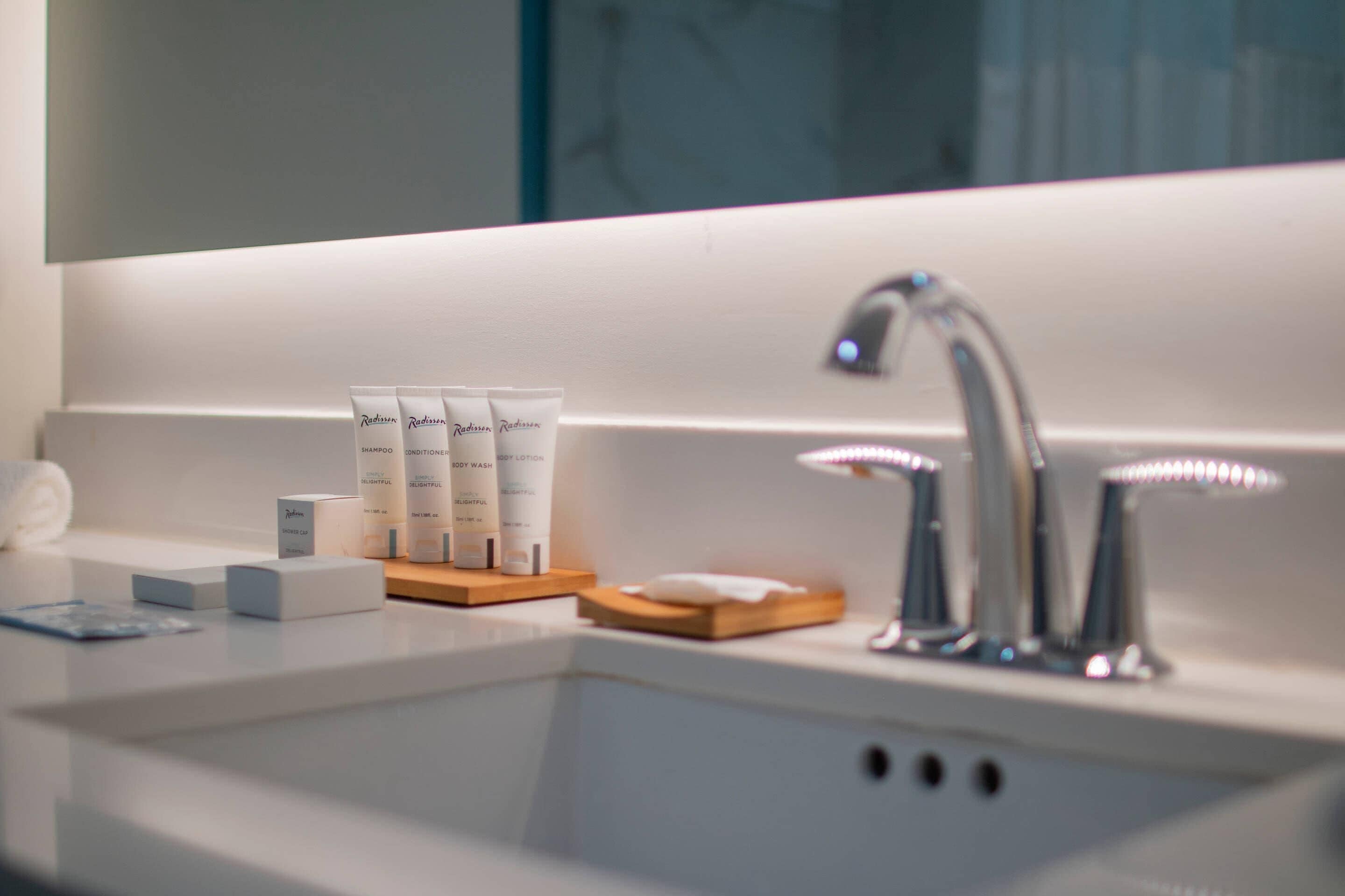 A bathroom with a white sink, two faucets, and a mirror above. There are three tubes of body wash and a box of shampoo on the sink.