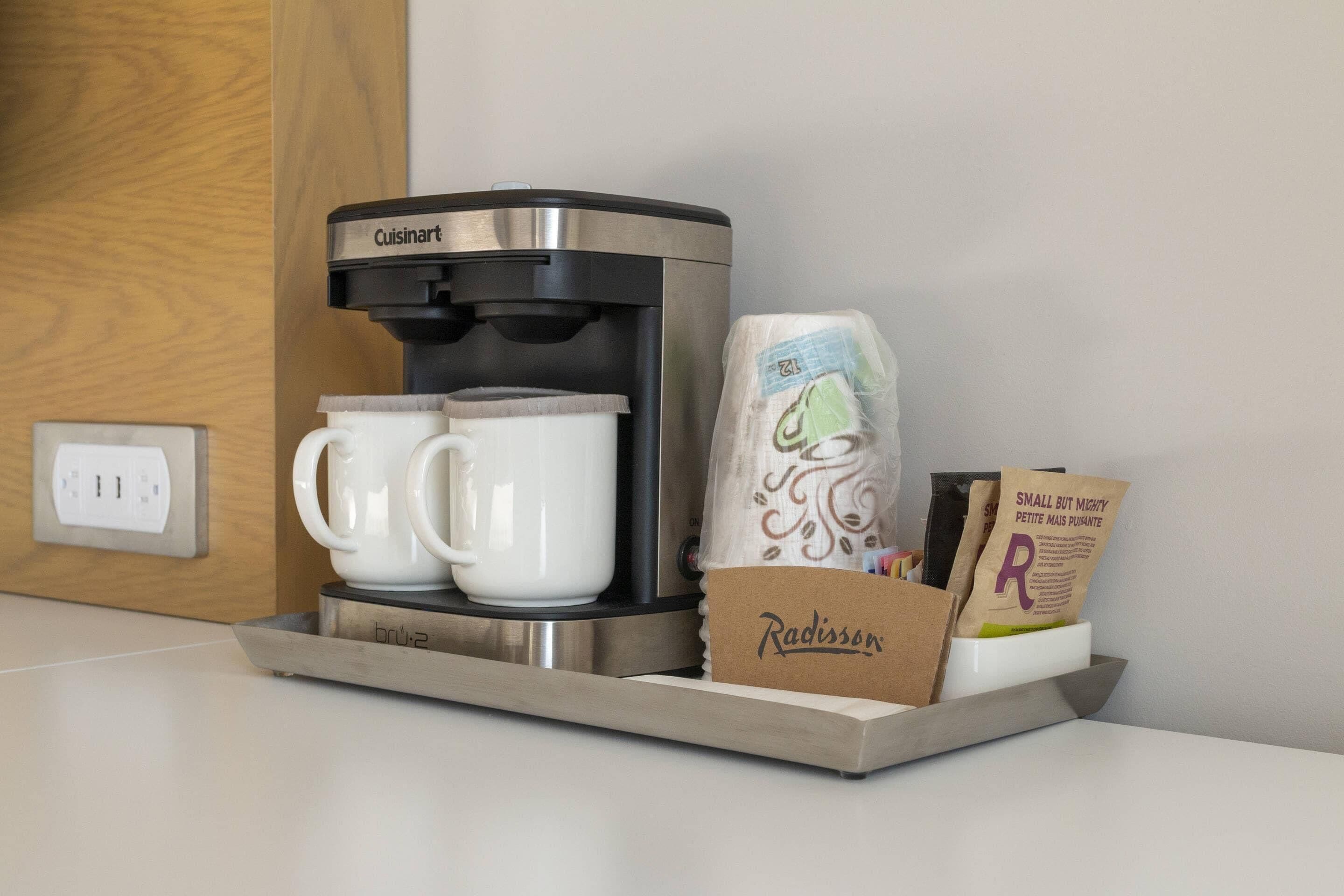 Une machine à café Cuisinart, deux tasses blanches, un sac en papier étiqueté « Radisson » et un porte-cartes avec la lettre « P ».