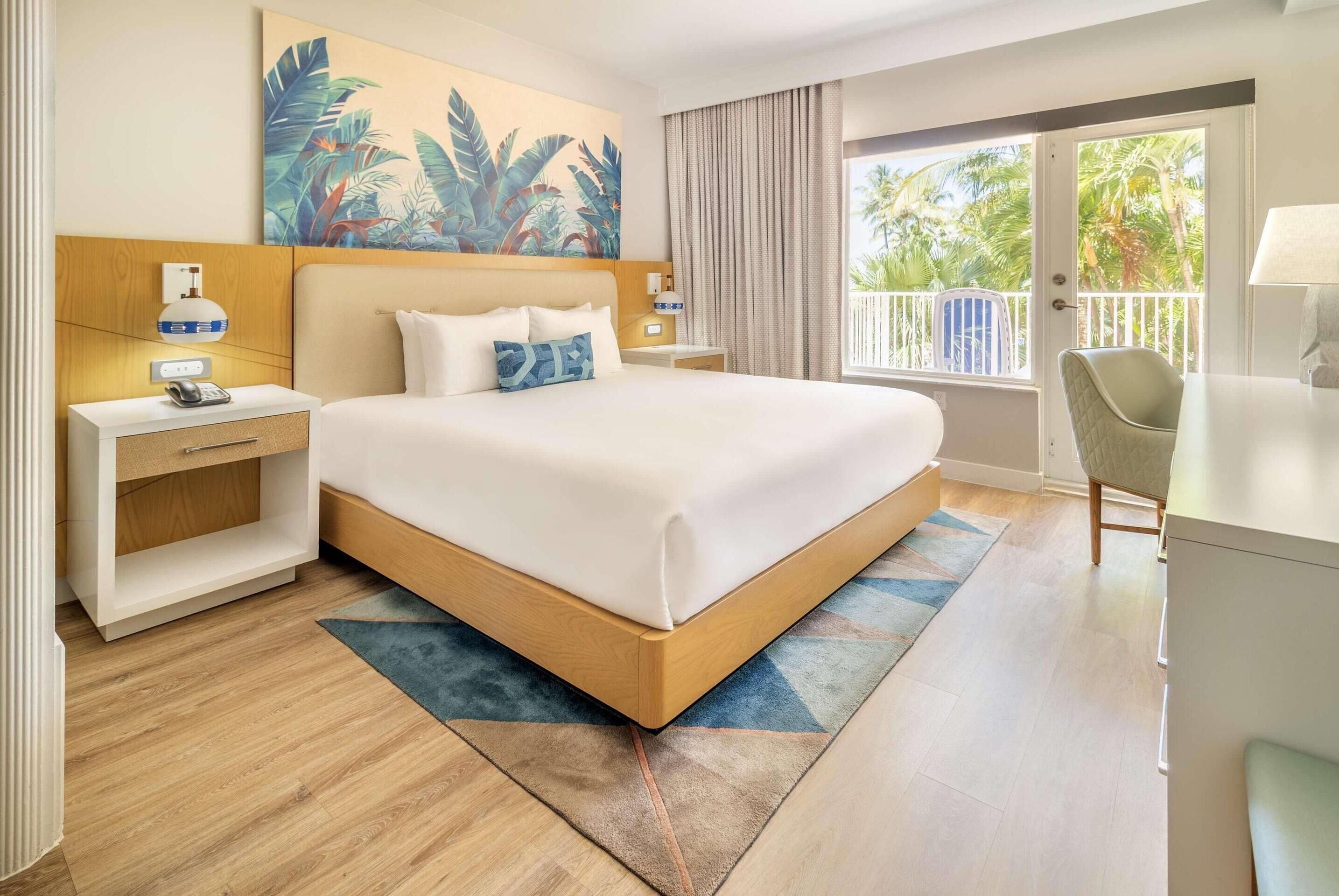 A hotel room with a large bed, a desk, a chair, a nightstand, and a painting on the wall.