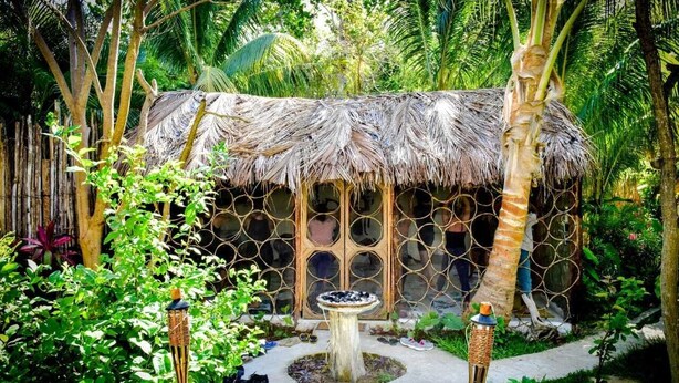 A thatched-roof structure with a circular window pattern, surrounded by lush greenery and a small circular water feature. - Casa Del Viento (Isla Holbox)