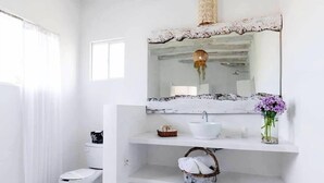 A white bathroom with a large mirror, a sink, and a toilet. There is a basket with towels and a vase with flowers on the sink shelf. - Casa Del Viento (Isla Holbox)