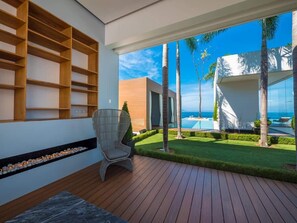 Un espace extérieur moderne doté d’une terrasse en bois, d’un coin salon avec un fauteuil, d’une étagère intégrée et d’une vue sur une piscine et des palmiers.