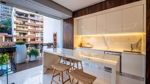 A modern kitchen with a marble countertop, wooden stools, and a view of a residential area.