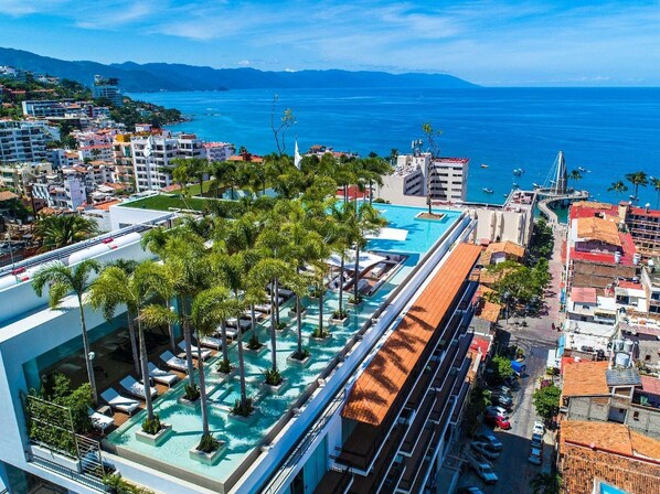 A rooftop pool area with palm trees, a view of the sea, and surrounding buildings.