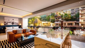 A modern hotel room with a large balcony, a glass coffee table, and a view of the surrounding buildings and greenery.