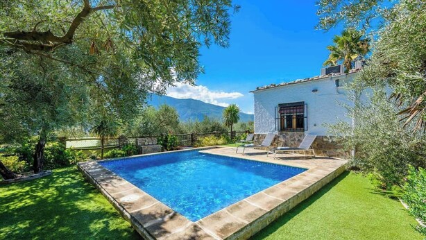 A rectangular swimming pool with a stone border, surrounded by a well-maintained lawn and trees. There is a white building with a window and a door to the right of the pool. - Las Gallinas Órgiva by Ruralidays (Granada)