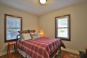 Un dormitorio con una cama, dos ventanas, una mesita de noche y una lámpara.