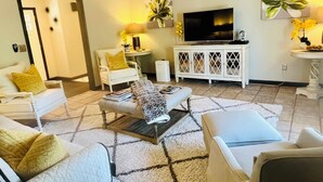 Una sala de estar con televisor, muebles blancos y plantas decorativas.