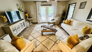 Una sala de estar con sofá, sillón, reposapiés y televisor. Hay una puerta con la leyenda 'FARMHOUSE' y un ventilador de techo.