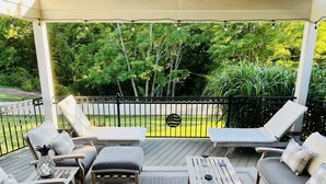 Un patio cubierto con mobiliario exterior, una mesa de madera y vistas a un césped y árboles bien cuidados.
