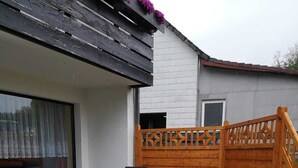 Een terras met twee loungestoelen en een kleine tafel, een houten schutting, en een huis met een witte gevel en een grijs dak.