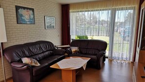 Een woonkamer met een donkerbruine leren bank, een bijpassende fauteuil en een kleine houten salontafel. Twee ingelijste foto’s hangen aan de muur, en er is een raam met gordijnen.