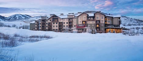 Un hôtel à plusieurs étages, avec un télésiège en premier plan, entouré de montagnes enneigées.