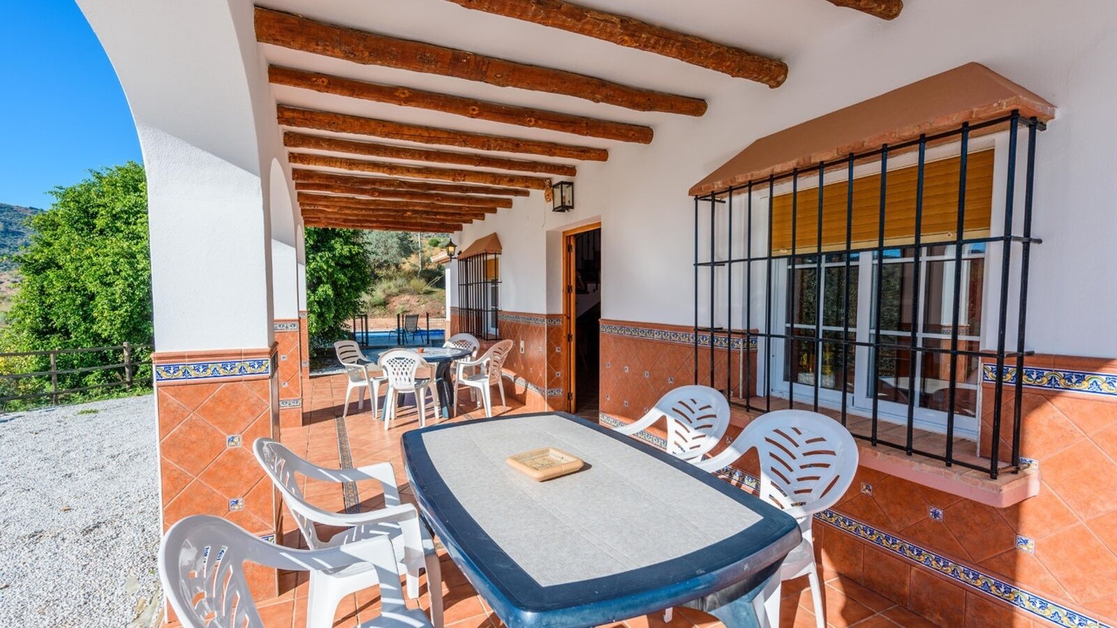 An outdoor patio with a table and chairs, a pool in the background, and a view of greenery.