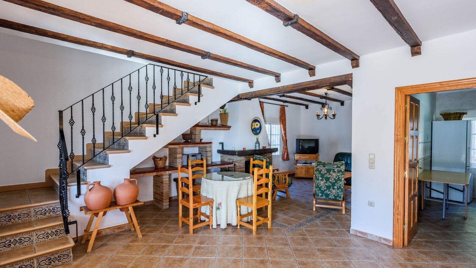 Un interior rústico con una escalera de madera, una zona de comedor con una mesa redonda y sillas, y una cocina con refrigerador y pileta.