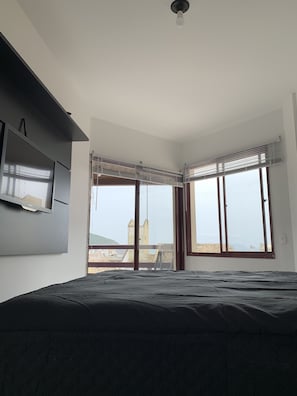 Un dormitorio con cama, televisión de pantalla plana en la pared y ventanas grandes con persianas.