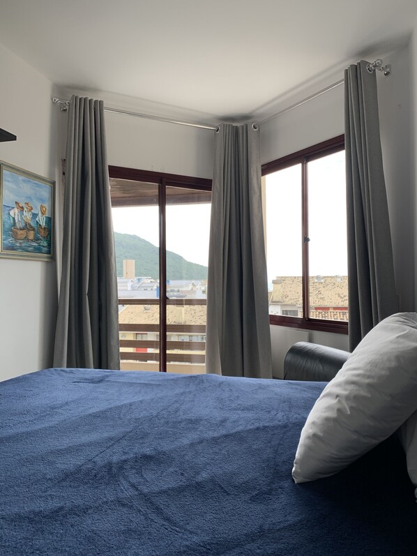 Habitación de hotel con cama, un cuadro en la pared y vistas a edificios y montañas a través de la ventana.
