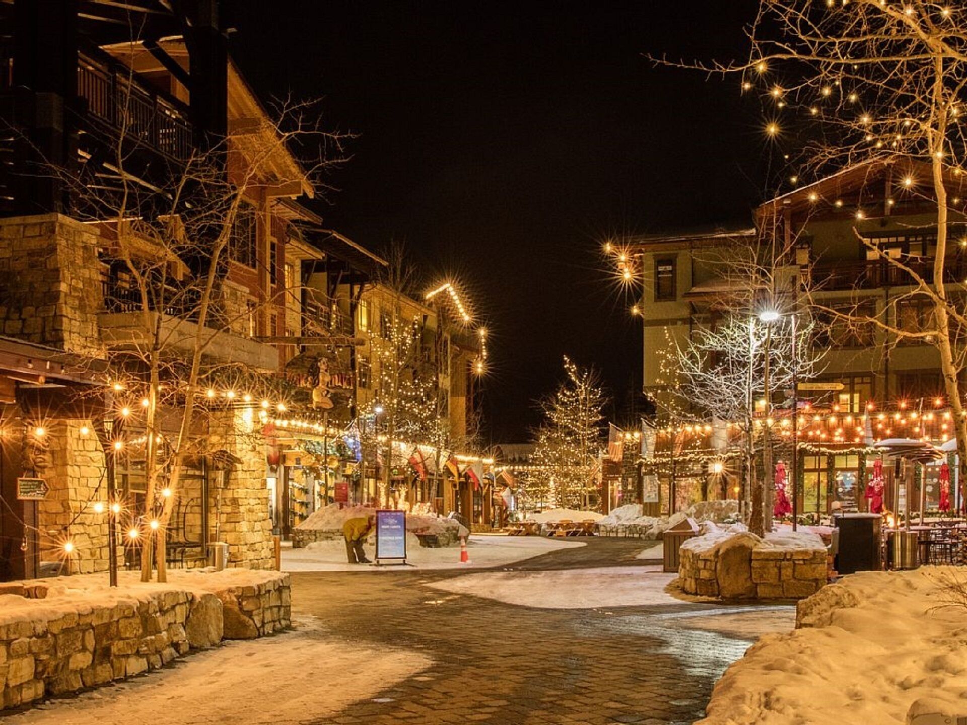 A snow-covered street with buildings adorned with string lights, a stone pathway, and a small stream.