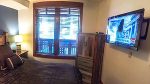 Habitación de hotel con cama, televisión, ventana con persianas y un cuadro en la pared.