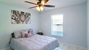 Une chambre à coucher avec un lit, un ventilateur de plafond, une fenêtre et une œuvre d’art représentant un tigre accrochée au mur.