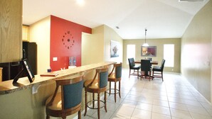 Une cuisine avec une horloge murale rouge, un coin bar avec des tabourets et un espace repas avec des chaises et une table.