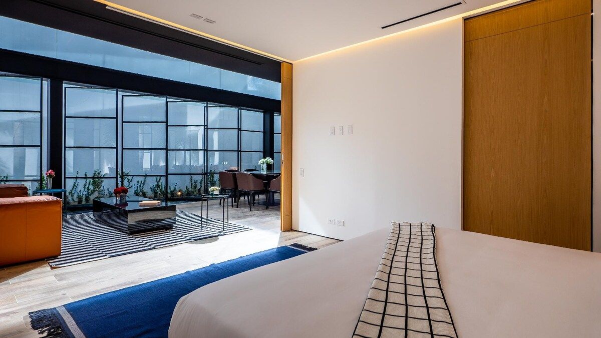 A modern hotel room with a large window, a bed, a seating area with a table, and a wooden wardrobe.