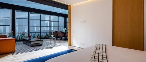 A modern hotel room with a large window, a bed, a seating area with a table, and a wooden wardrobe.