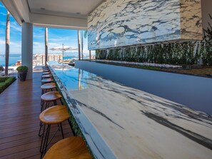 A poolside bar with a marble countertop and wooden stools, overlooking a body of water with a cityscape in the background.