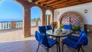 An outdoor patio with a round glass table, blue chairs, and a decorative wall with a patterned arch. - Villa el Pedregal Frigiliana by Ruralidays (Málaga)