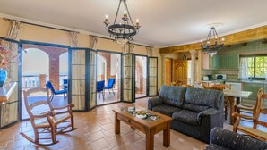 A spacious living room with a sofa, chairs, and a coffee table. There is a balcony with blue chairs and a view of the outside. The room has a chandelier and a kitchen area visible in the background. - Villa el Pedregal Frigiliana by Ruralidays (Málaga)